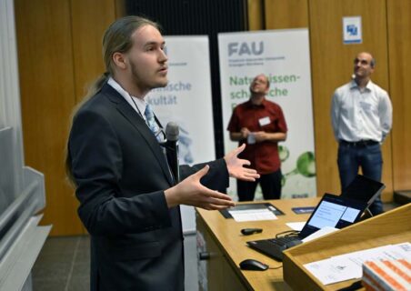 Arvid Traue von der Beruflichen Oberschule der Stadt Nürnberg, 1. Platz Mathematik, beim Vortrag. Im Hintergrund sind links Dr. Johannes Hild (FAU) und rechts Prof. Dr. Julien Bachmann (FAU) zu sehen. (Foto: Harald Sippel / FAU)